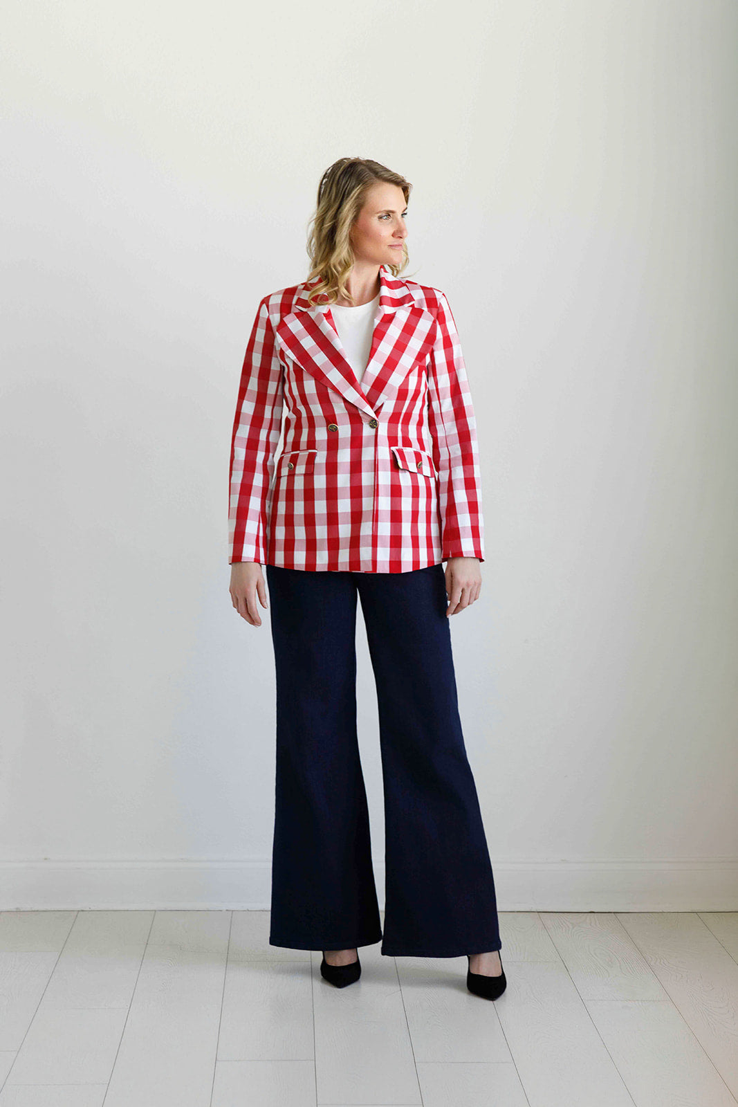 A woman stands in a studio wearing a red and white gingham blazer over a white top, paired with dark navy wide-leg pants and black heels.