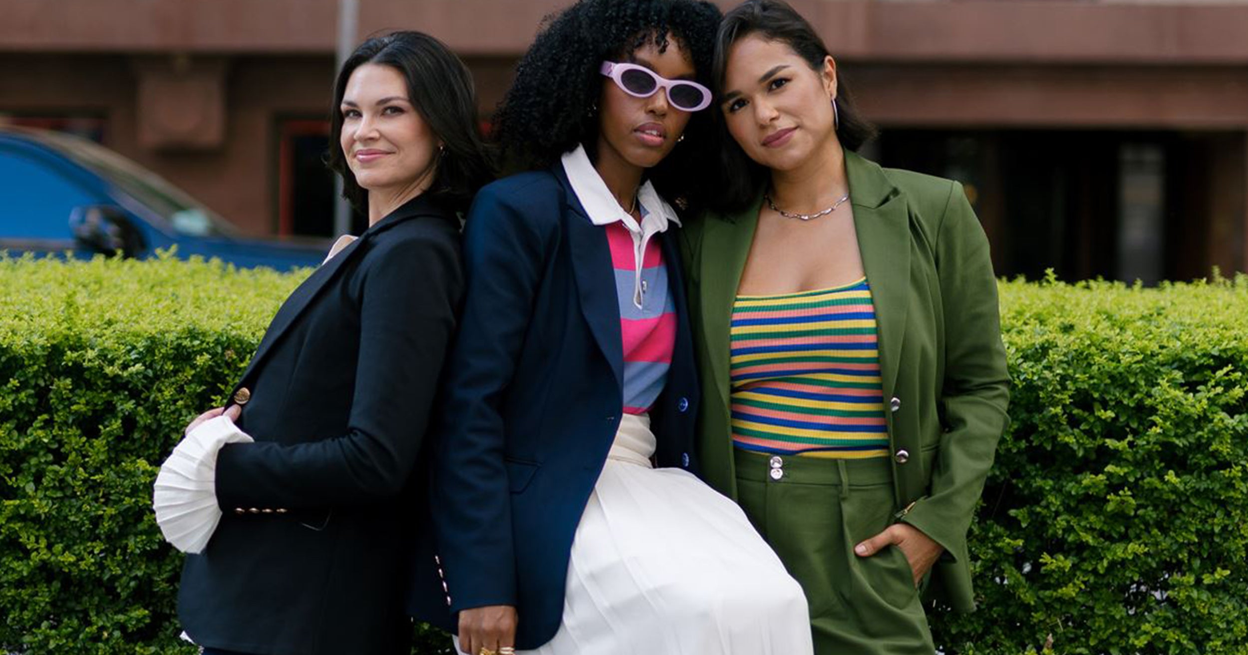 Three women posing together outdoors in front of a hedge. The woman on the left wears a black blazer and white gloves.