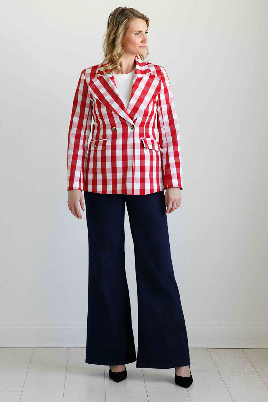 A woman stands in a studio wearing a red and white gingham blazer over a white top, paired with dark navy wide-leg pants and black heels.
