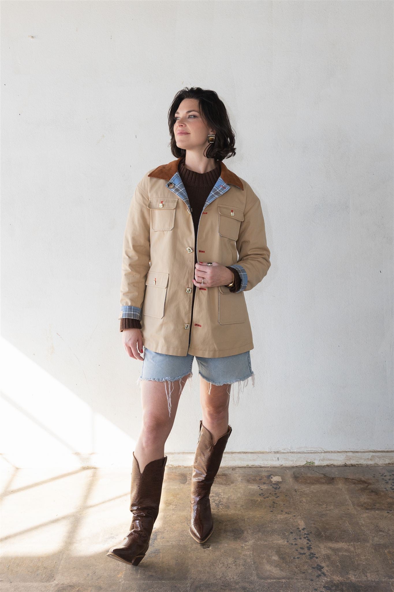 Woman wearing a beige barn jacket with brown trim, denim shorts, and brown boots against a white wall. power woman