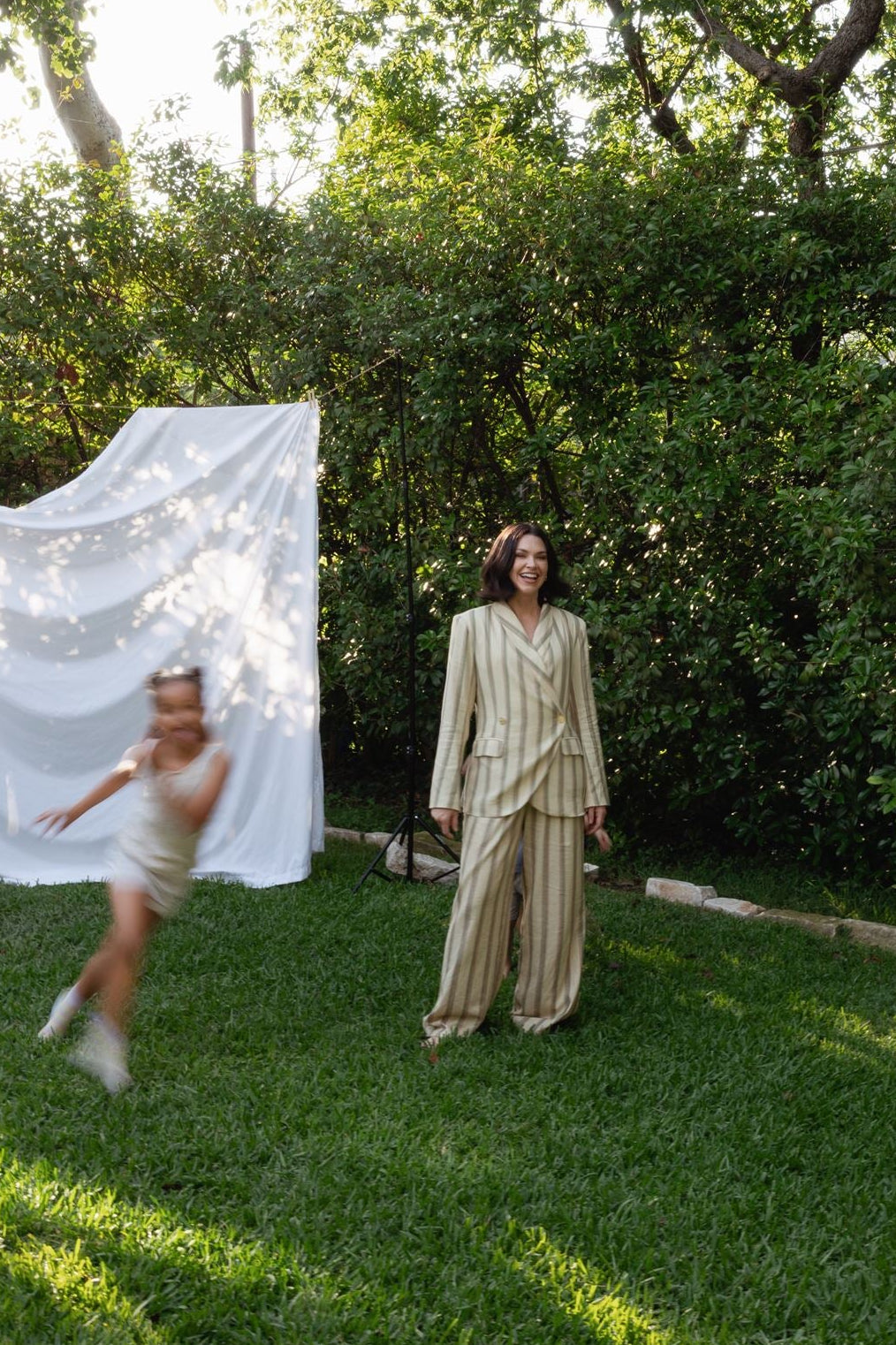 A woman in a beige matching two-piece suit stands on a grassy lawn in a garden, with a child blurred in motion on the left near a white draped fabric backdrop. Trees and greenery fill the background.