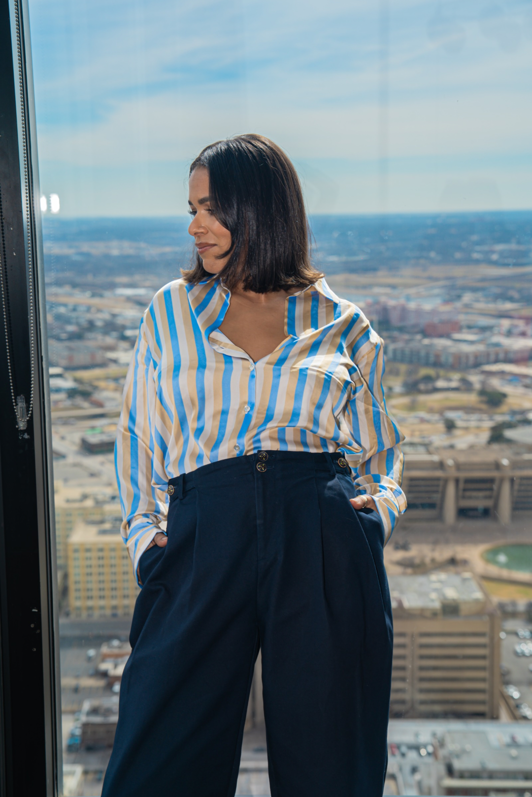 Woman in a striped shirt and high-waisted pants standing in front of a large window with a cityscape view. womens trouser pwr wmn power woman 
