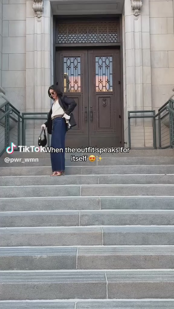 Woman poses on a stone staircase wearing PWR WMN’s Miss Noir Black Oversized Blazer with dark denim and heels.