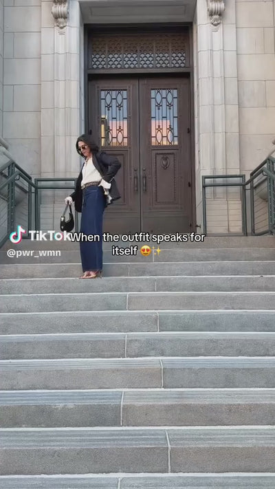 Woman poses on a stone staircase wearing PWR WMN’s Miss Noir Black Oversized Blazer with dark denim and heels.